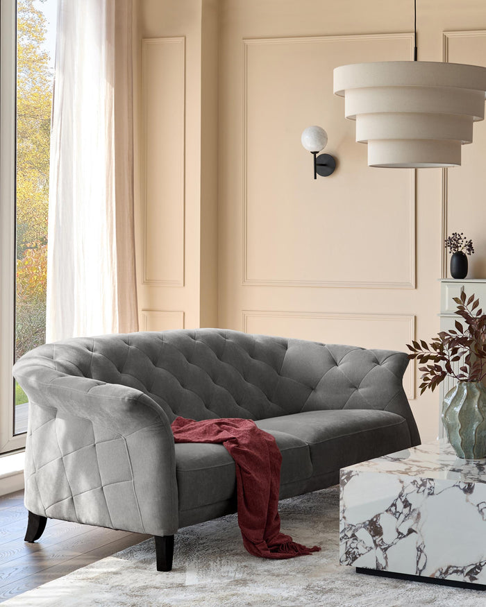A grey, buttoned sofa sits in a neutrally decorated room with a marble coffee table and hanging lamp.