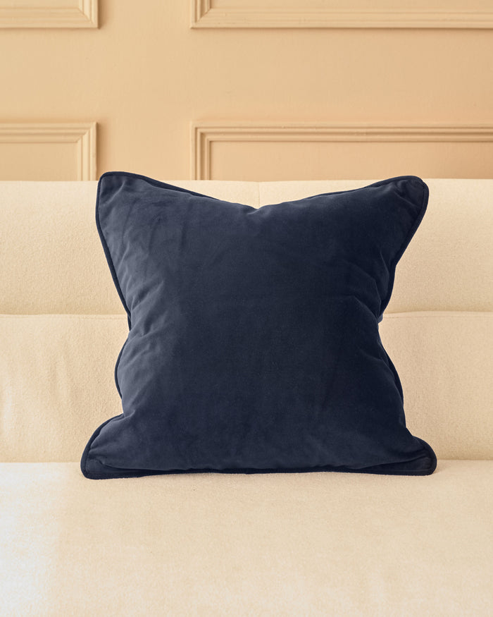Navy velvet cushion with piping detail, displayed on a cream upholstered sofa against a beige paneled wall.