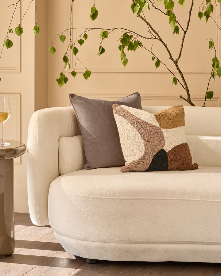 Lennon cushion in natural displayed on a cream curved armchair with geometric patterned pillows in a styled living room.