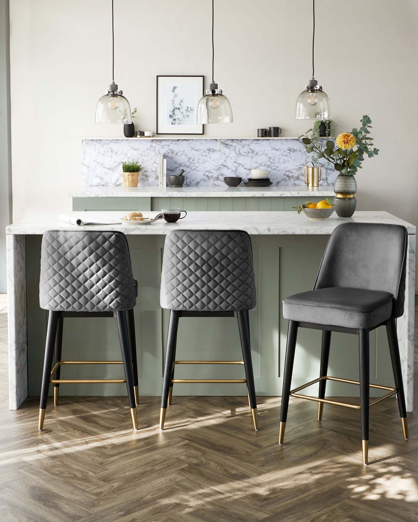 Elegant contemporary bar stools with grey quilted velvet upholstery and black legs accented with gold footrests. The bar stool design features a sleek silhouette and a high backrest, paired with a white marble kitchen island.