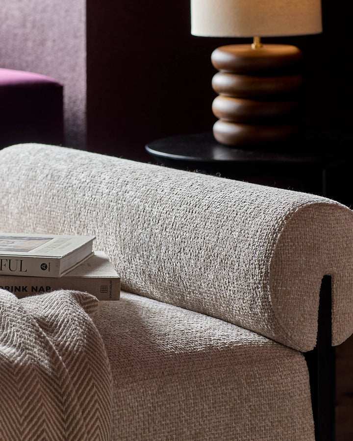 Maeve daybed in natural weave fabric, shown at an angle with rolled backrest and stacked books detail.