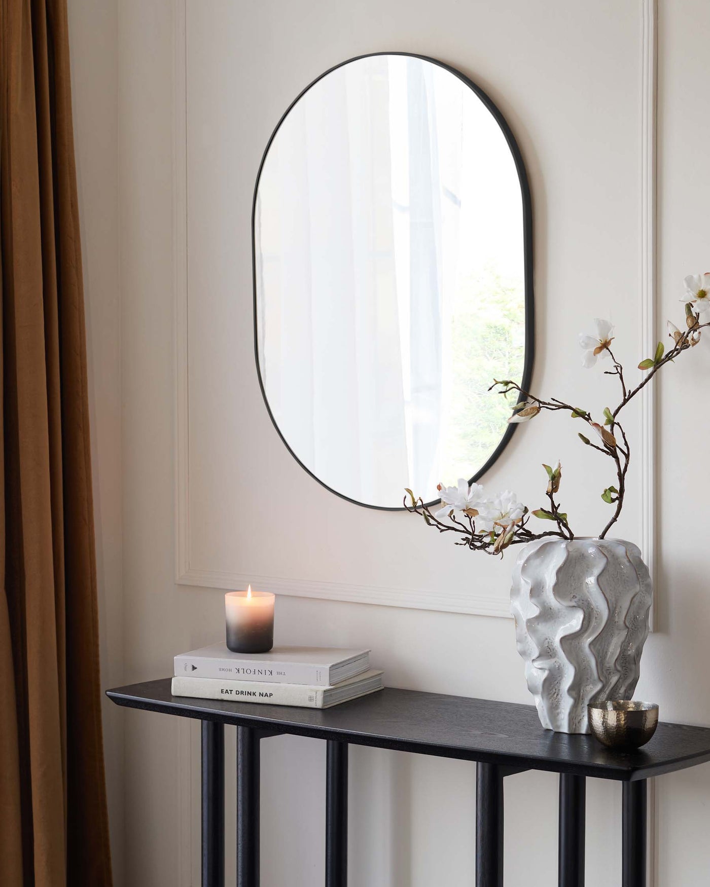 A contemporary console table with a dark grey, textured surface, and a minimalist black metal frame featuring straight lines and right angles. The table is accessorized with books, a lit candle, and a sculptural white vase with flowering branches. A round mirror with a thin black frame hangs above the table against a neutral wall, enhancing the modern aesthetic.