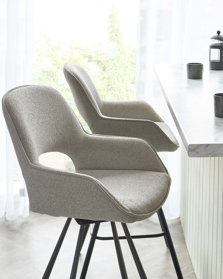 Two modern light grey upholstered bar chairs with mid-height backs, curved armrests, and slender black metal legs are situated near a white marble countertop. The chairs feature a textured fabric and are positioned in a bright, airy space with sheer white curtains partially revealing lush greenery outside.
