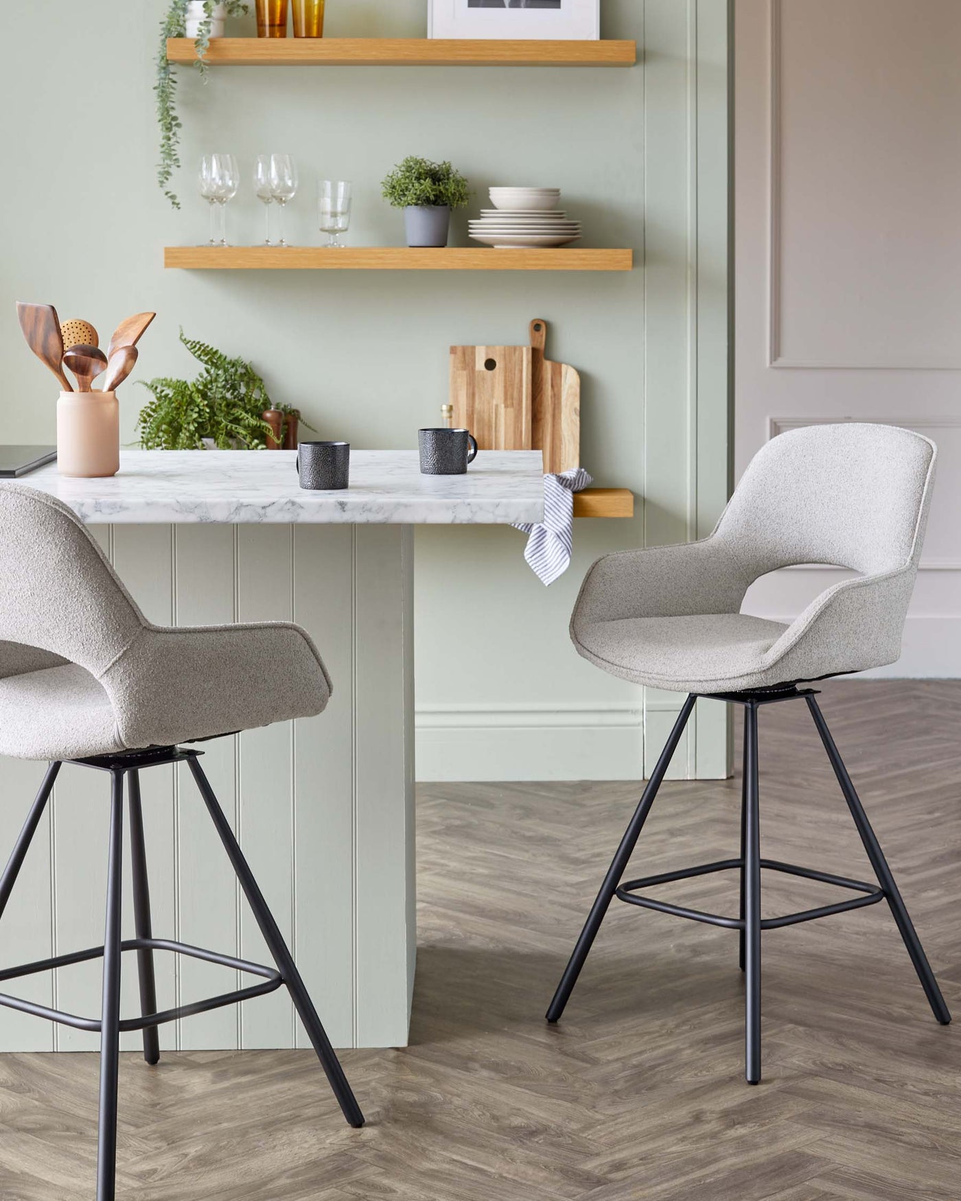 Two modern, fabric-upholstered, bar-height chairs with a curved backrest and slim, black metal legs beside a white marble-topped kitchen island with pale green cabinetry. Wooden shelves with dinnerware and plant accents in the background.