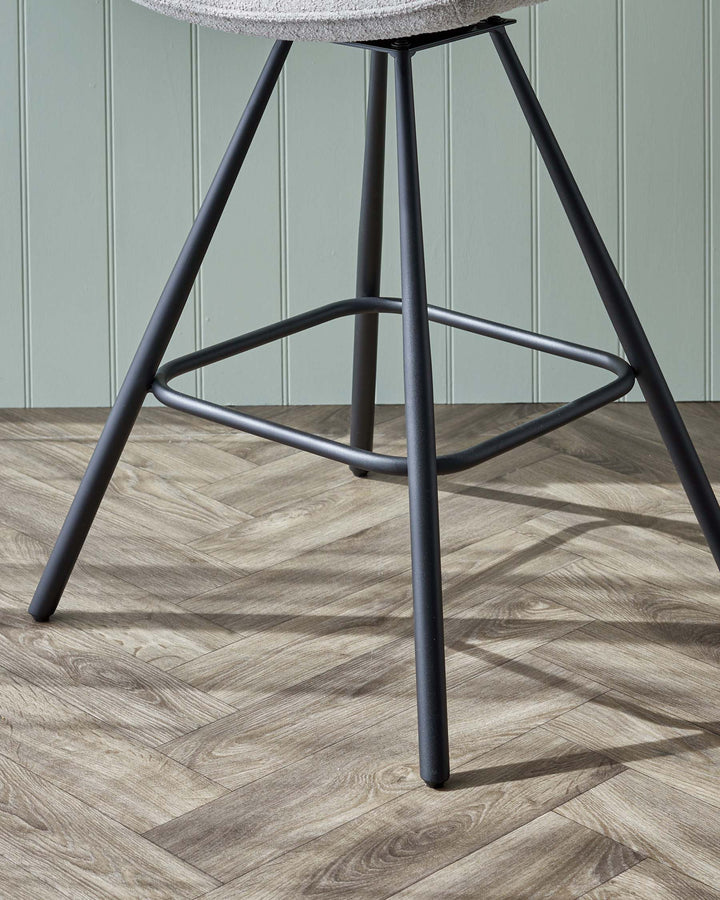 Modern stool with textured light grey upholstered seat and black metal legs on a herringbone pattern wooden floor.