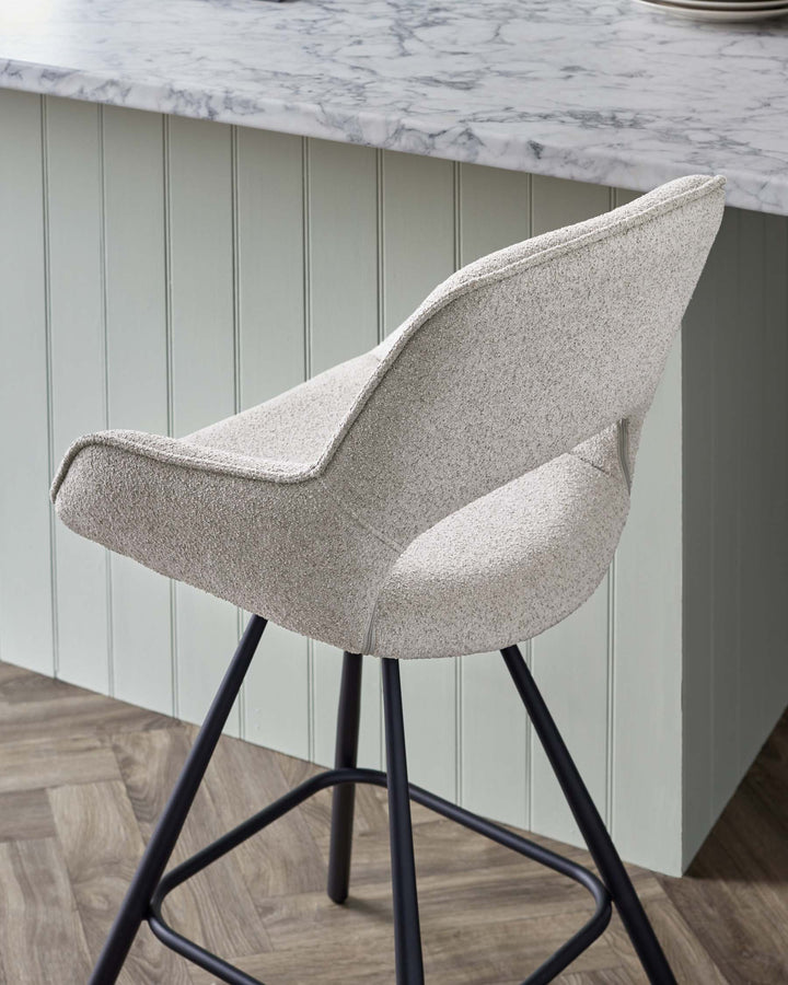 Modern upholstered bar stool with a textured light grey fabric and wingback design, featuring a supportive backrest and a well-cushioned seat. The stool stands on slender, black metal legs with a geometric base for stability. The contemporary style complements the marble countertop and pale green panelled bar backdrop.