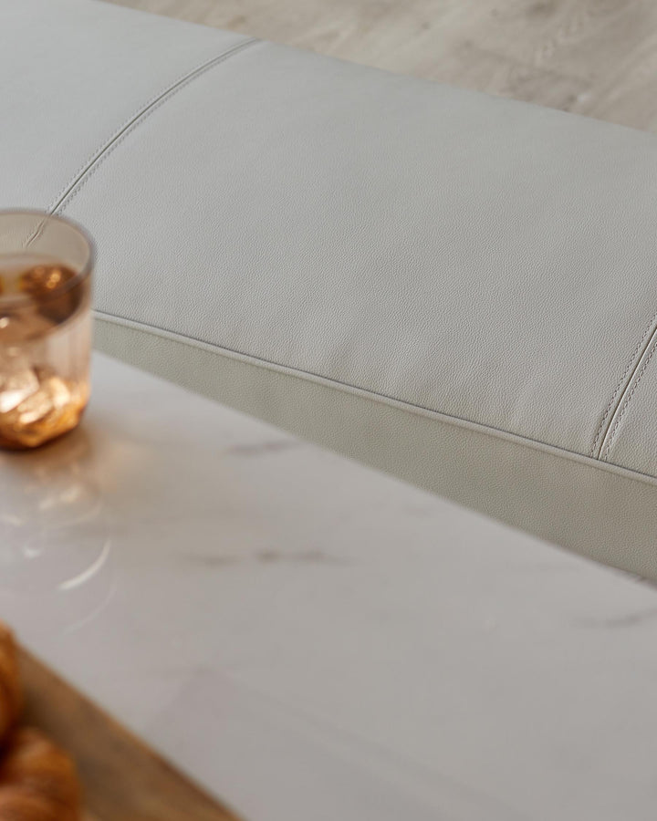 Close-up of a stylish grey leather sofa with stitching details next to a marble table with a glass and pastries.