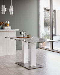 A rectangular, glass-topped dining table with two white legs and a metal base, positioned in a bright, modern dining area.