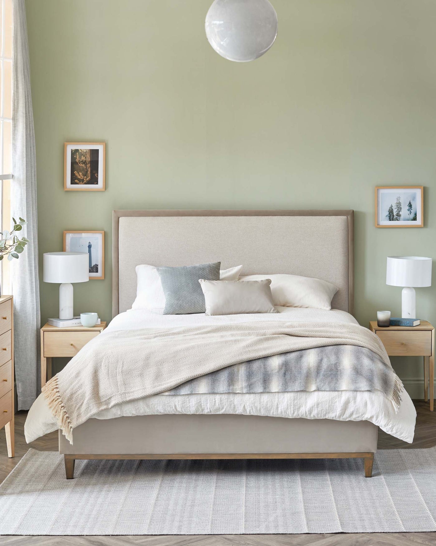 Contemporary bedroom furniture set featuring a minimalist upholstered king-size bed with a neutral-toned headboard and wooden legs, flanked by two light wood nightstands with clean lines and white table lamps. A soft beige area rug under the bed adds a layer of texture to the serene space.