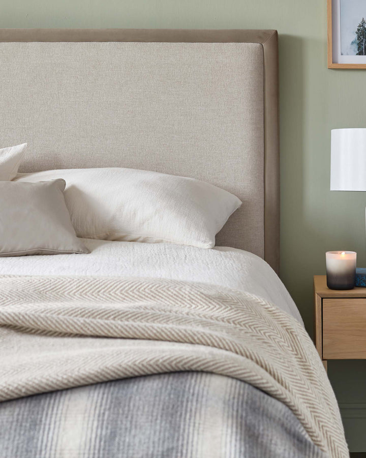 A neutral bedroom with a linen headboard, crisp white bedding and a textured throw. A wooden bedside table features.