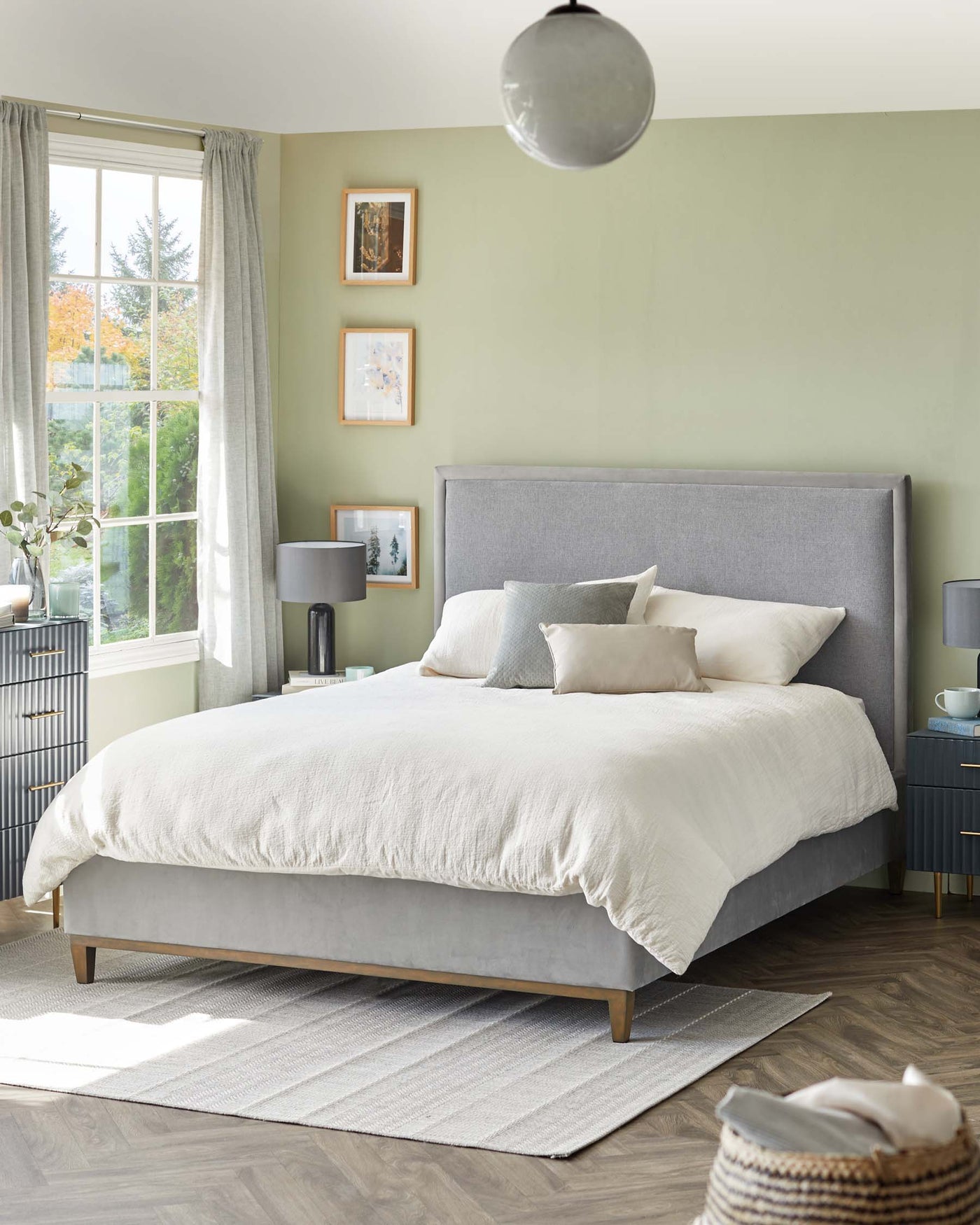 A modern bedroom featuring a grey upholstered platform bed with a high headboard and wooden legs, dressed with white and light grey bedding. A pair of round-top blue nightstands with gold handles flank the bed. The room has a large grey area rug under the bed and a woven basket on the floor to the right. A sleek black table lamp with a cylindrical shade sits atop one nightstand.
