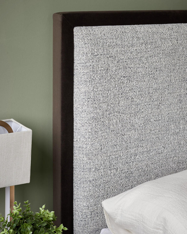 Upholstered headboard in grey fabric with a dark brown trim, on a bed with white bedding, with a lamp on the side table.