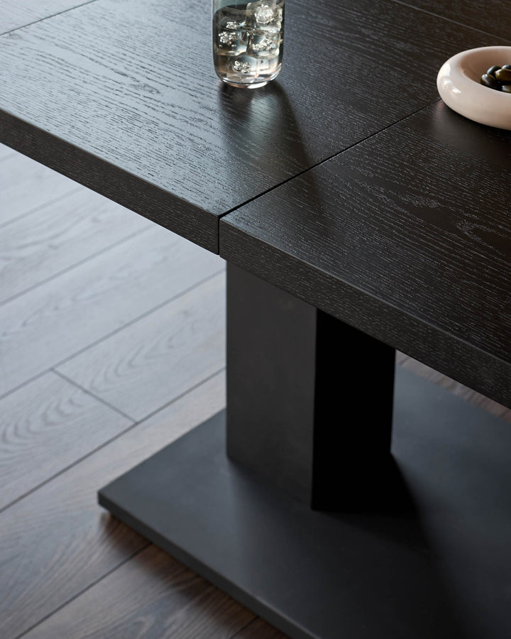 Dark wood dining table with textured finish, shown at angle displaying sleek pedestal base and tabletop detail.