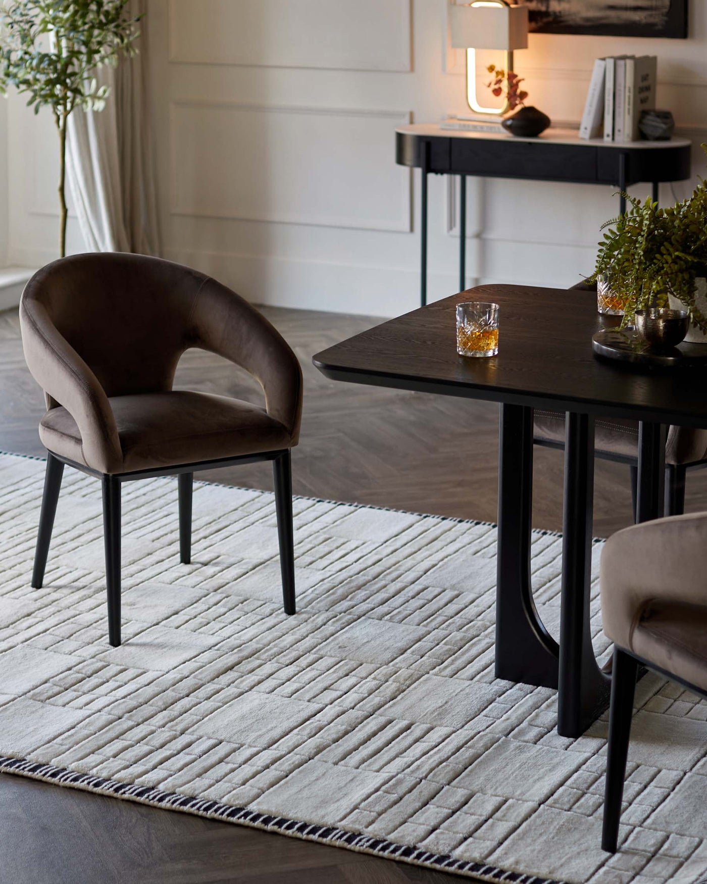 A contemporary dining room featuring a sleek, dark wood rectangular table with black metal legs, complemented by plush, curved velvet dining chairs in a muted taupe colour also with black legs. The arrangement sits atop a textured off-white rug with geometric lines and a black border, creating a sophisticated and modern space. A minimalist console table with decor items is partially visible in the background.