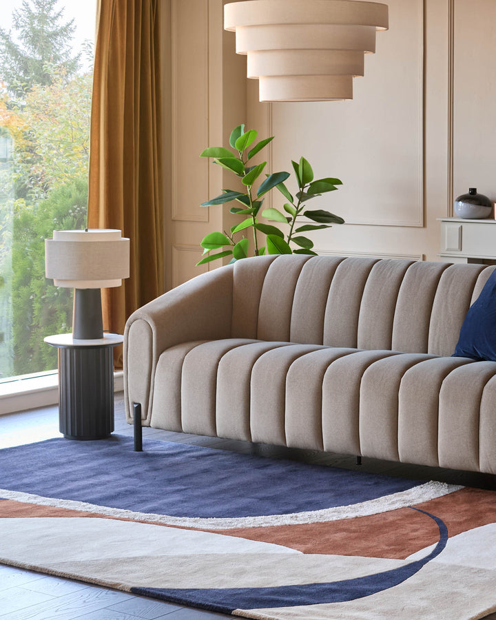 Leonie table light in linen natural with mocha base, displayed in a modern living room setting beside a curved sofa.
