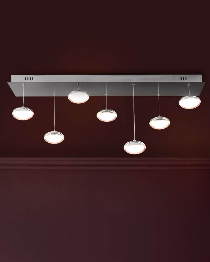 Seven round glass pendant lights with chrome fixtures suspended from a rectangular chrome ceiling plate against a deep burgundy wall.