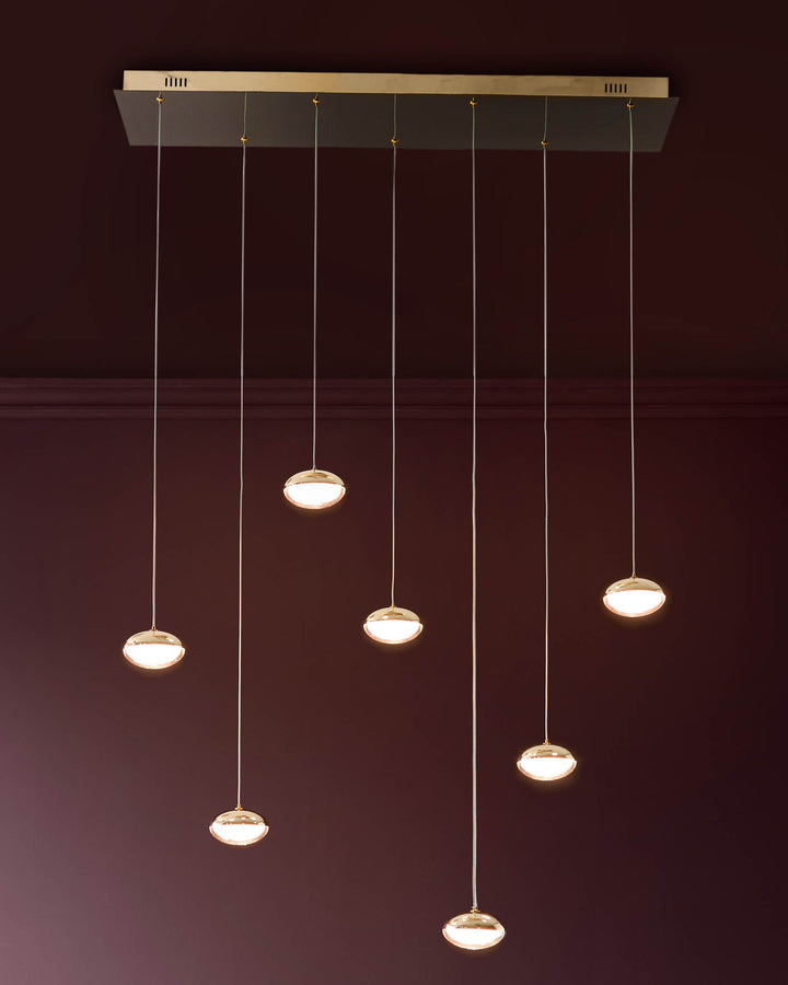 Nina 7-pendant light in soft gold finish with glass shades, suspended from rectangular canopy against burgundy wall.