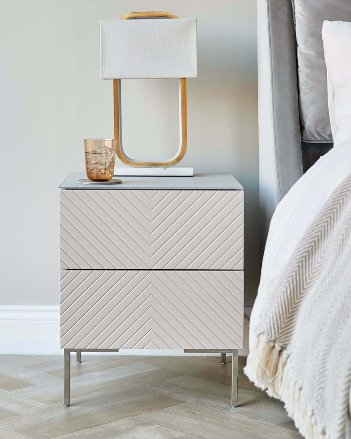 Stone Noah bedside table with chevron pattern drawers, metal legs, topped with wooden-framed lamp and glass of water.