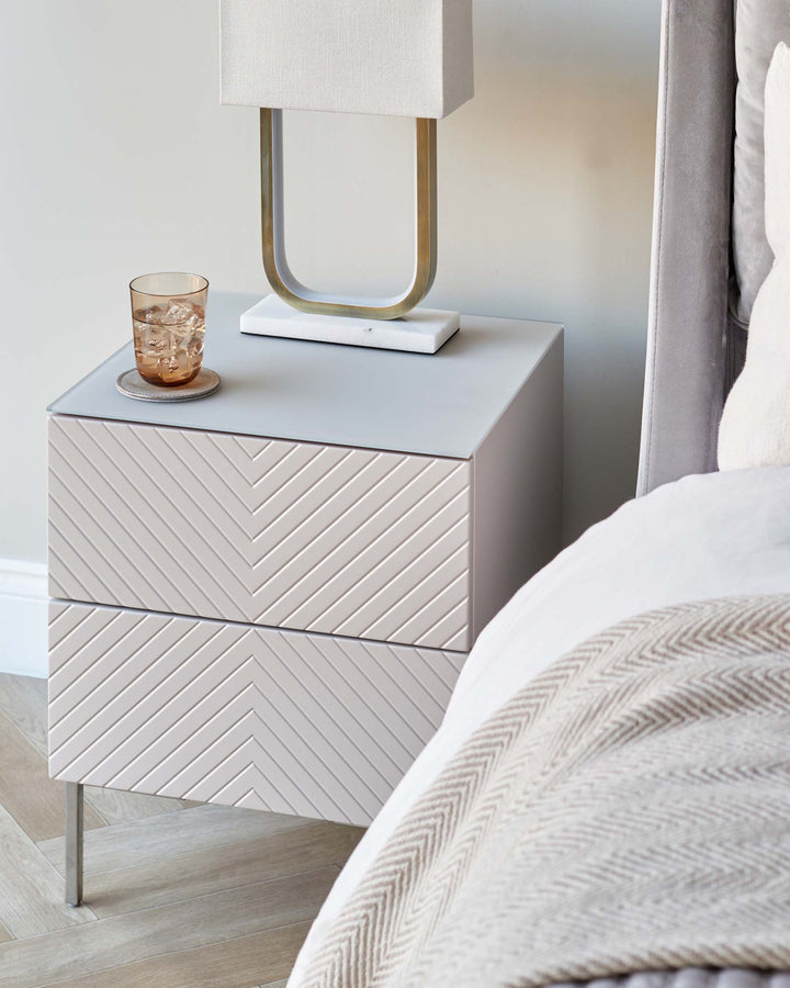Modern bedside table with chevron design and a glass of water, next to a bed with a grey headboard.
