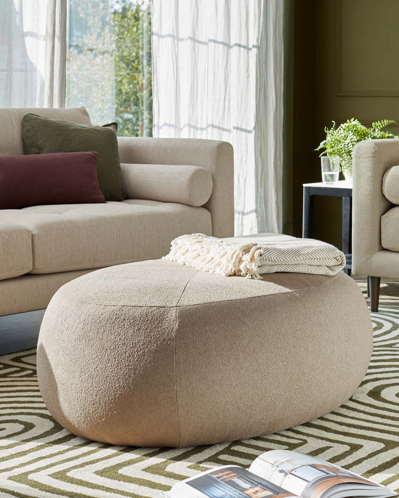 A modern beige upholstered armchair with rounded contours and a matching oversized round ottoman in a plush fabric. The ottoman is adorned with a cream-colored textured throw blanket. A small dark grey side table with a potted fern and glass cup is visible in the background. The setting is completed with a geometric-patterned area rug in complementary neutral tones.