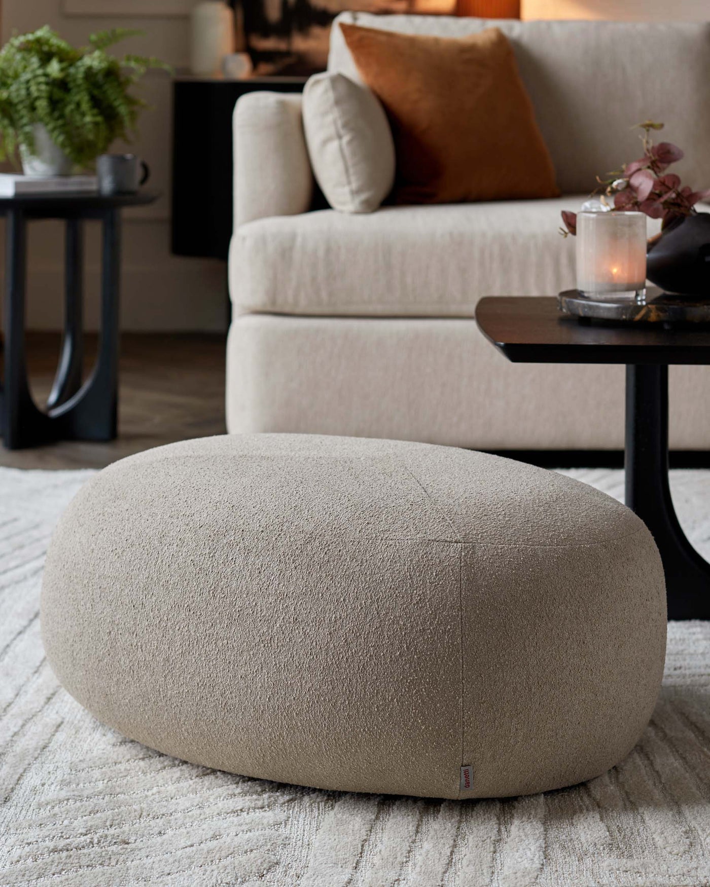 Beige textured oval-shaped ottoman on a white area rug with a beige upholstered sofa, black round side table with a candle, and small green potted plant in a cosy home interior setting.
