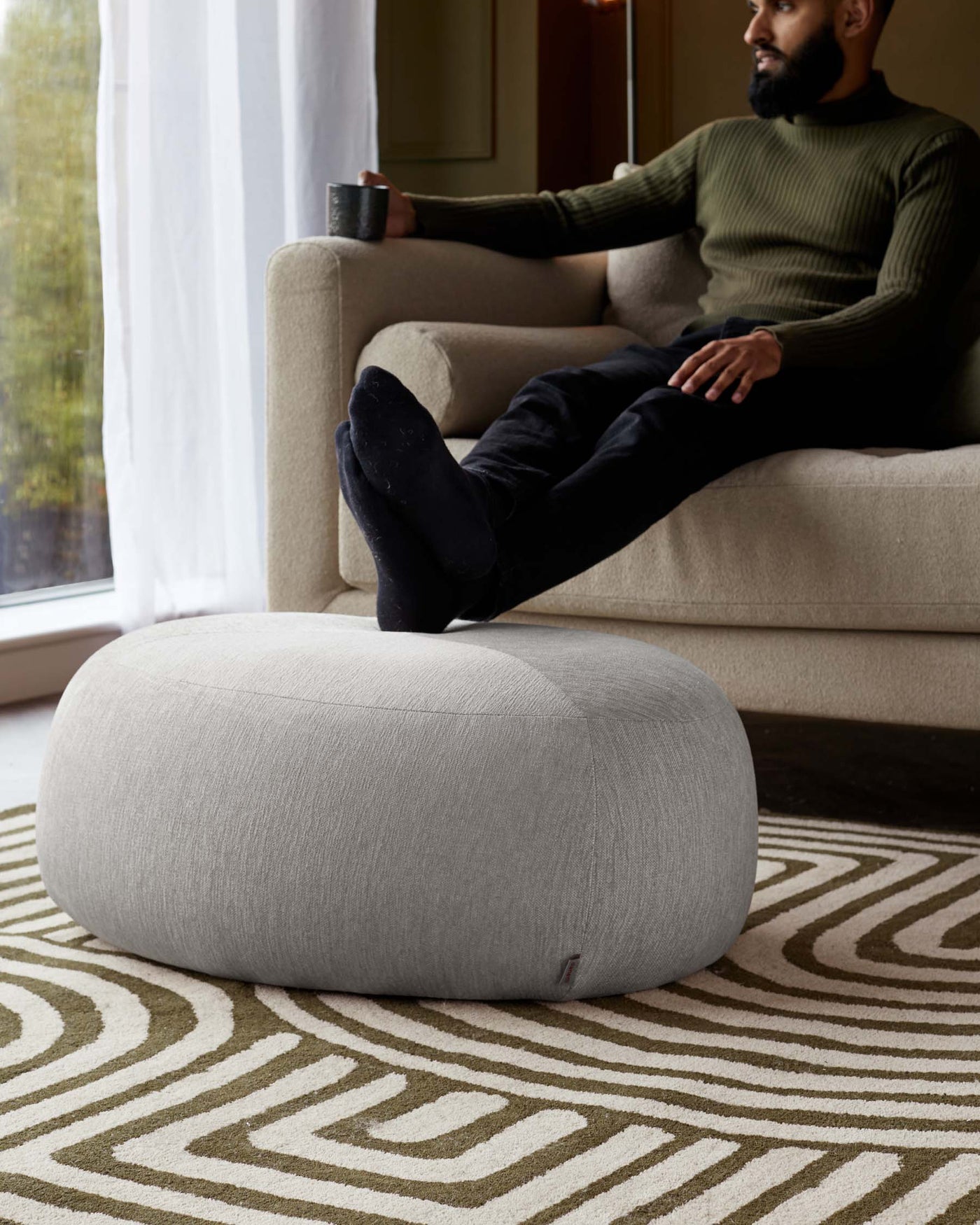 A neutral-toned modern fabric ottoman with a smooth, rounded design resting on a geometric patterned area rug in shades of beige and light brown. A beige contemporary sofa with clean lines is partially visible to complement the ottoman.