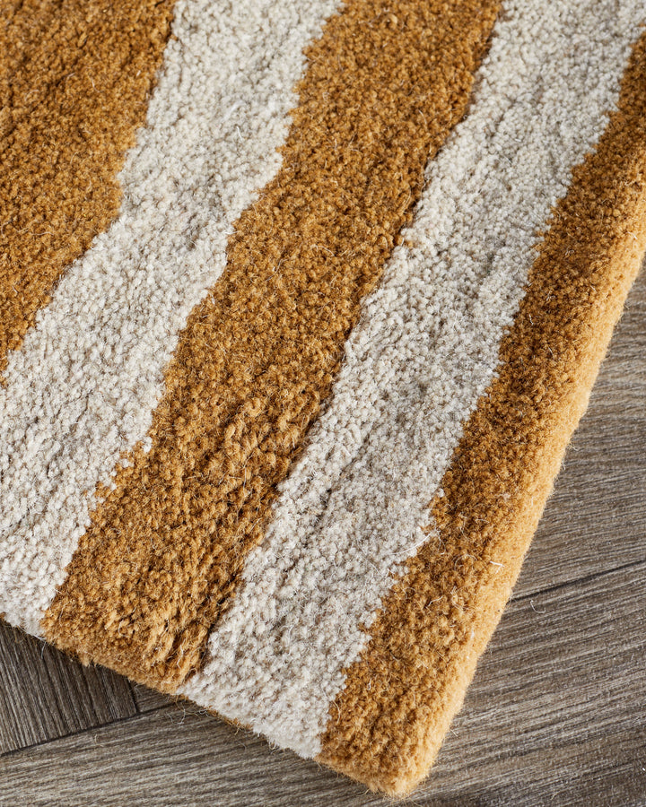 Small sculpted wool rug with abstract swirl pattern in ochre and cream, shown at an angle on a wood floor.