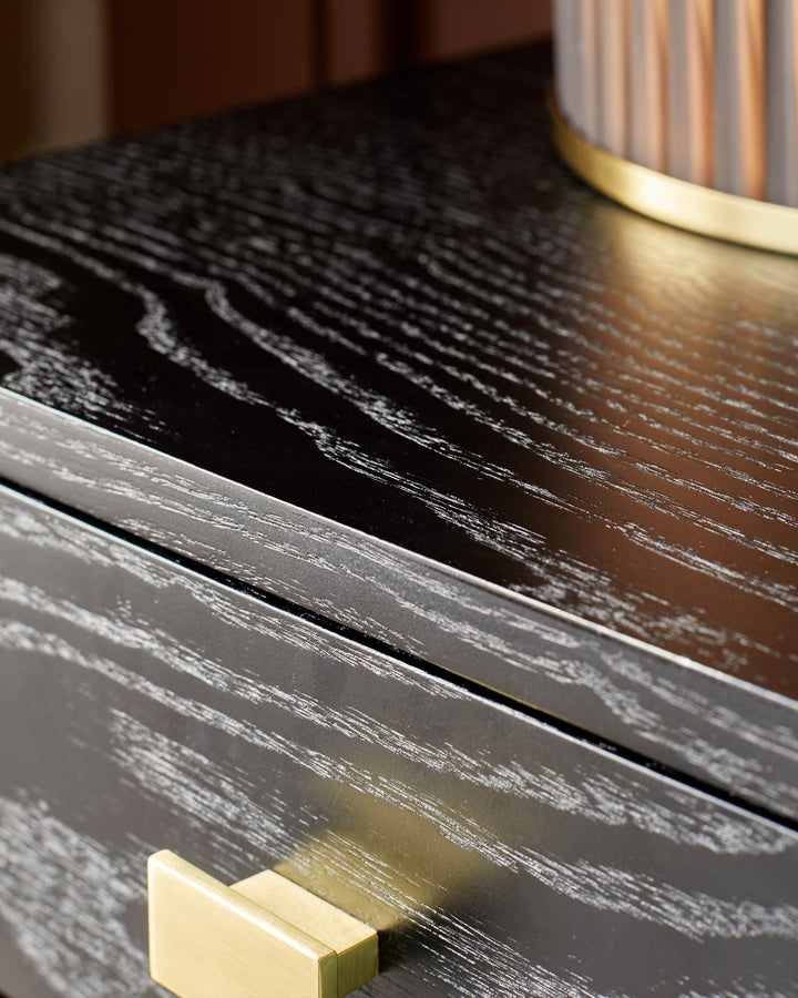 Close-up of a black wood grain bedside table, featuring a brass drawer handle and stylish lamp.