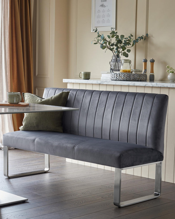 A grey upholstered dining bench with brushed metal legs sits adjacent to a glass table.