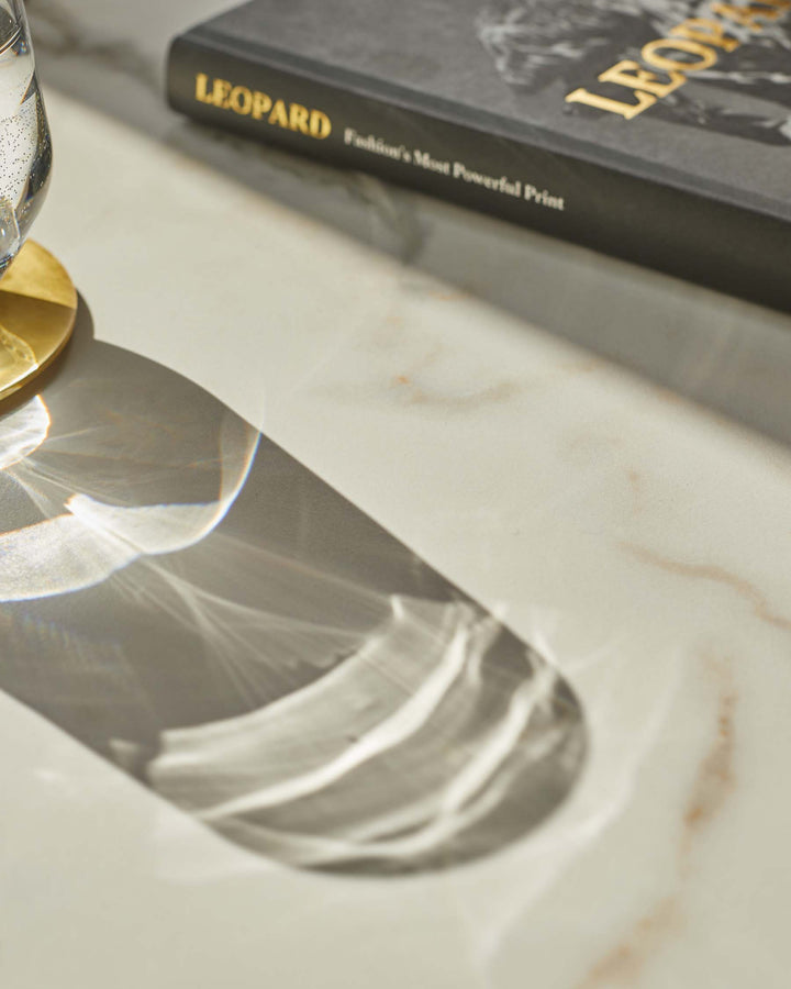 A marble-patterned table featuring the shadow from a glass, a gold coaster, and a book titled 'Leopard, Fashion's Most Powerful Print'.