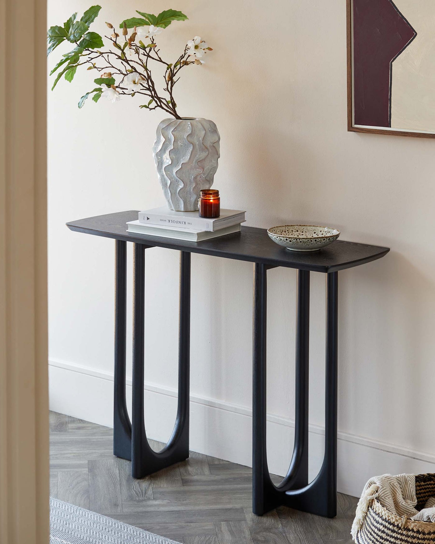 A modern, narrow console table with a dark, wood-grain finish top and a unique, sculptural base comprising of two curved, matte black metal legs.