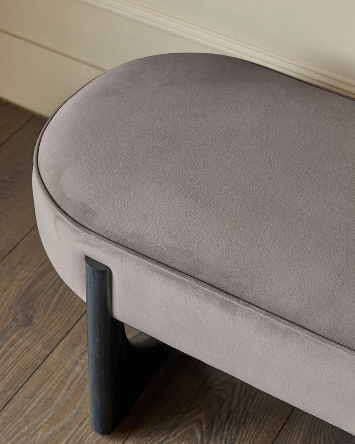 A stylish taupe velvet ottoman seat with dark wood legs sits on a wooden floor.