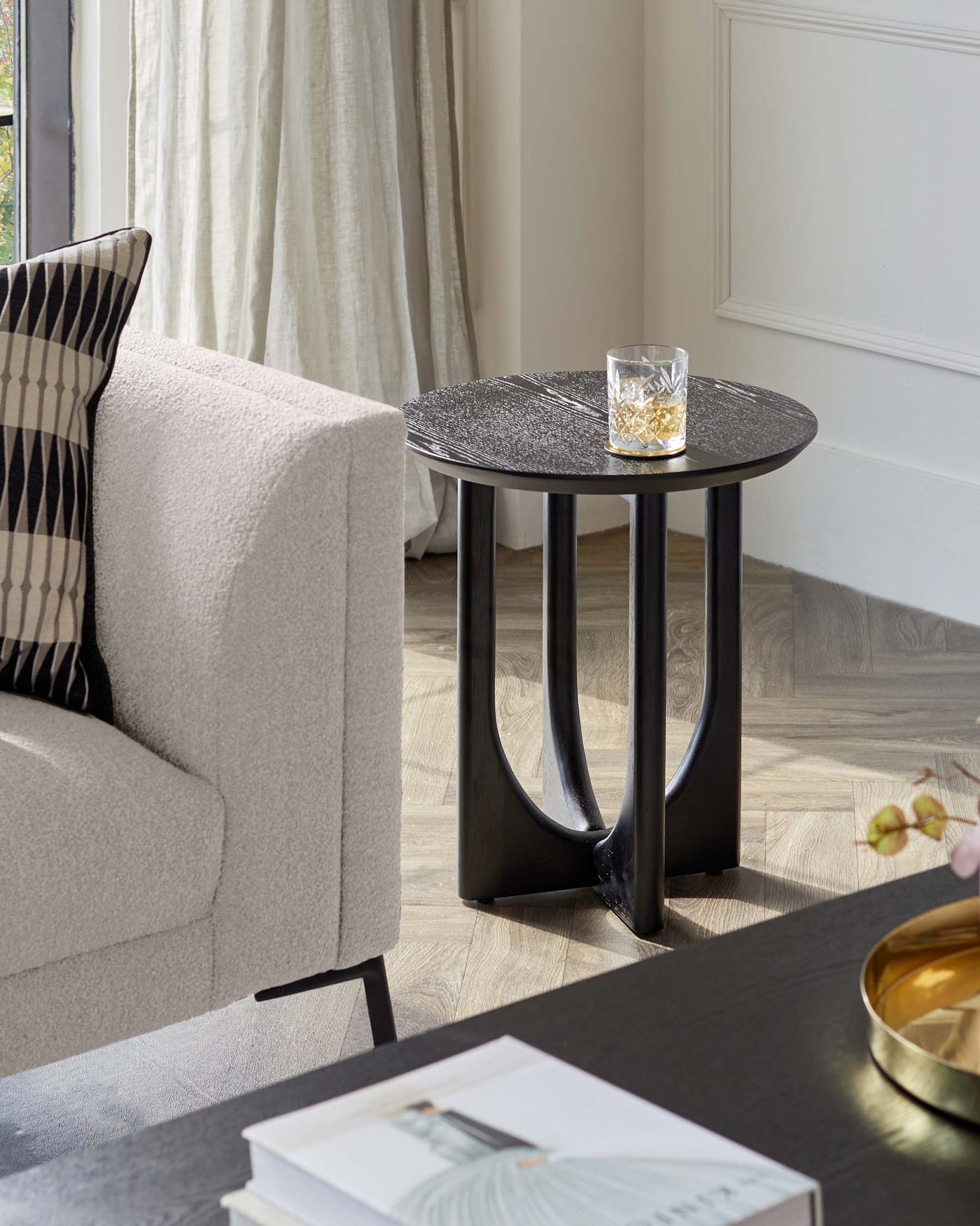 A contemporary round side table with a textured black tabletop and curved black metal legs. It is accessorized with a clear glass containing a beverage.