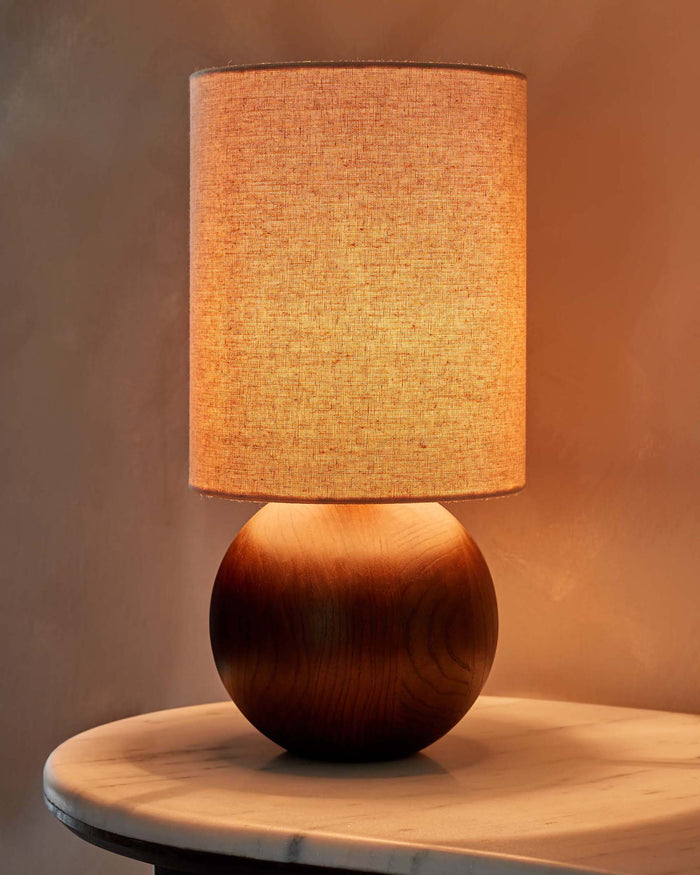 A stylish bedside lamp with a round wooden base and linen shade sits on a marble side table, providing a warm glow.