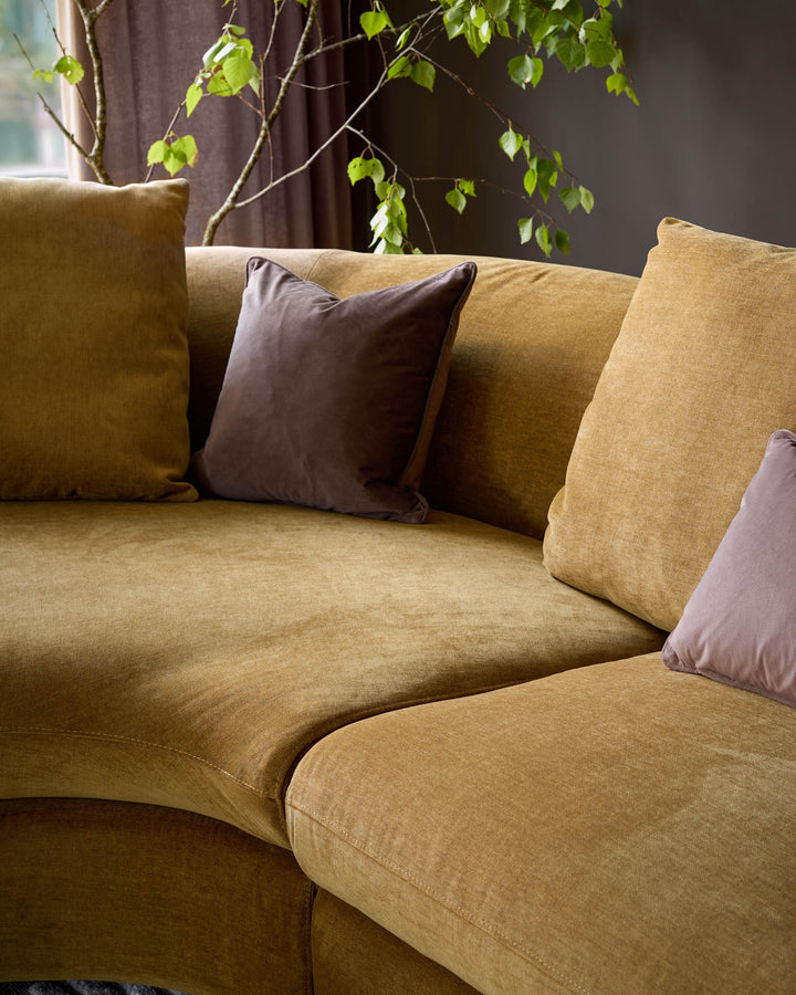 Ochre left-hand corner chaise sofa with mauve cushions, angled view showing curved seating and textured fabric finish.