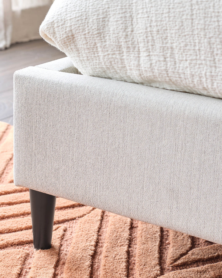 Close up of a cream coloured sofa with a textured cream cushion and a dark wood leg on a patterned salmon rug.