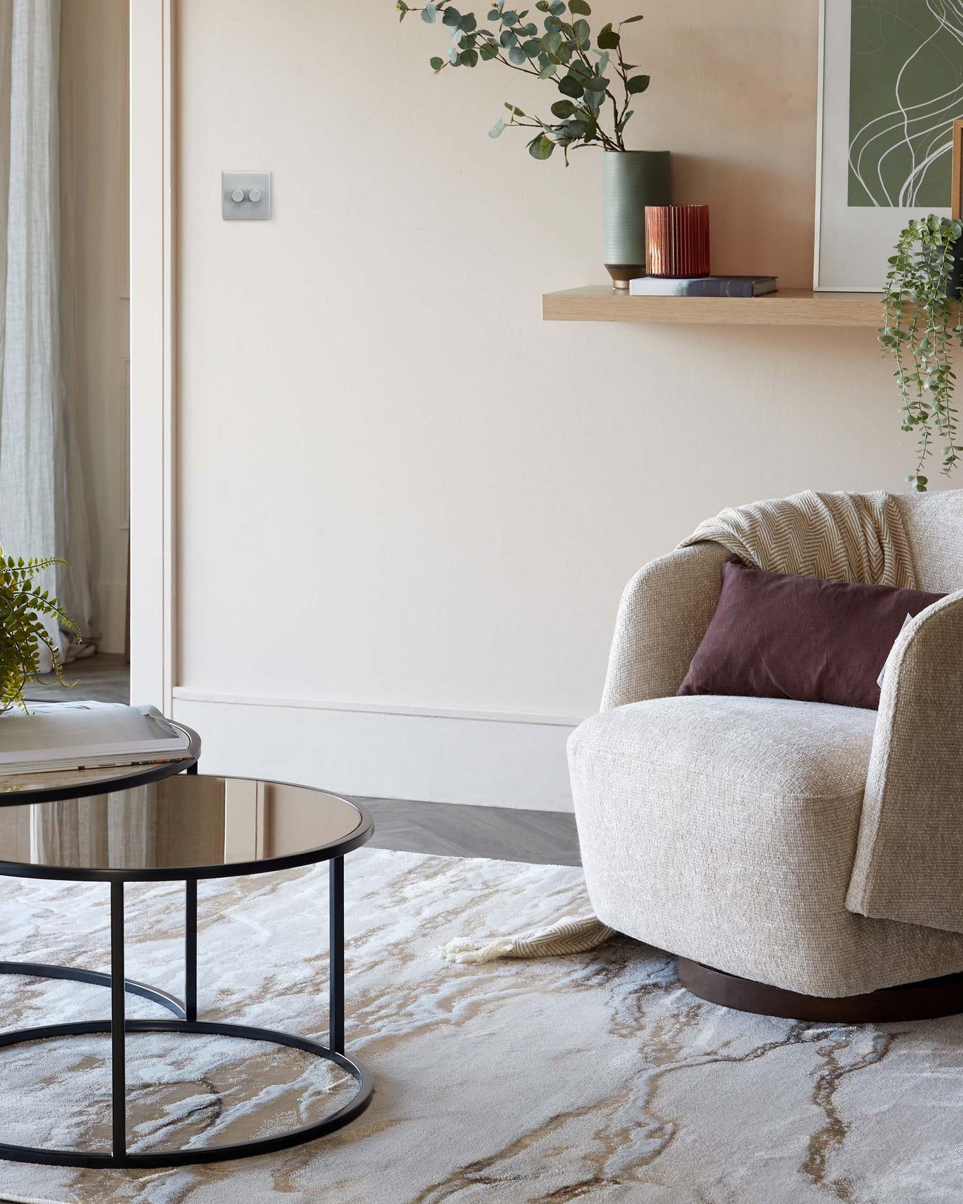 Modern living room featuring a round, black-framed glass-top coffee table and a cosy beige upholstered armchair with a textured throw pillow. An ornamental floating wooden shelf is mounted above, adorned with potted greenery and decorative objects.