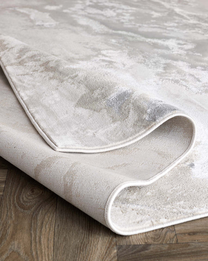 Close-up of a cream rug with a textured pattern and a rolled-up corner, revealing its underside and the wood floor beneath.