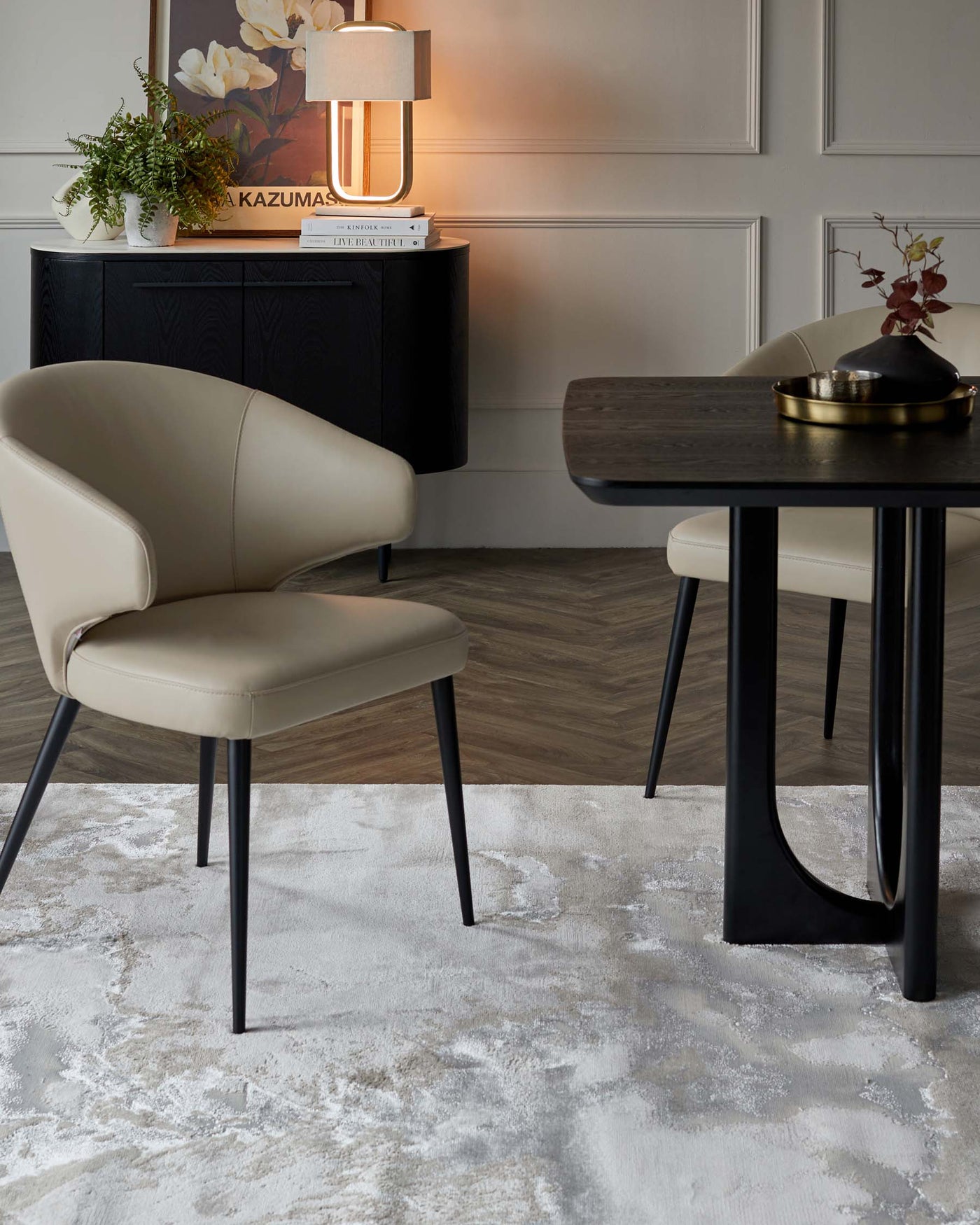 Elegant modern furniture set featuring a beige upholstered chair with a curved back and black tapered legs, a matching dark wooden table with black legs, and a textured black sideboard against a panelled wall, complemented by a white patterned area rug. A contemporary lamp, decorative books, and a brass tray with accessories adorn the sideboard, adding a sophisticated touch to the decor.