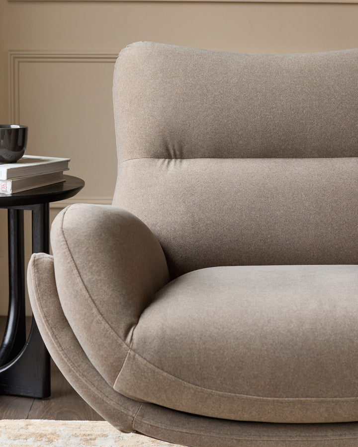 Mocha fabric accent chair with curved design, shown at angle with matching footstool beside black side table.