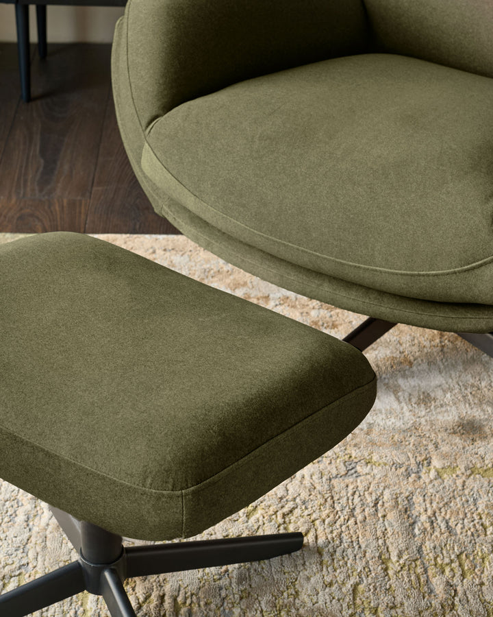 Piper accent chair with footstool in olive green velvet, shown from a side angle on a cream textured rug with dark wood flooring visible.
