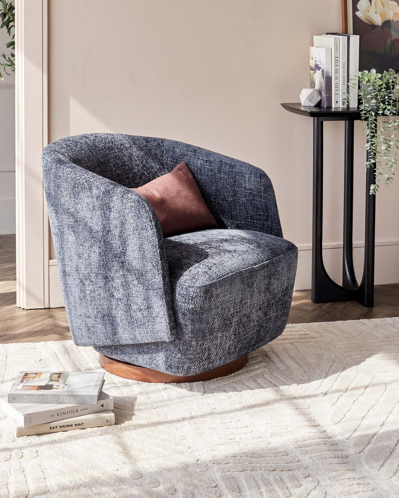 Contemporary rounded armchair with textured blue upholstery and a smooth wooden base, paired with a modern black circular side table with slender legs, holding books and decorative items, positioned on a neutral-toned textured rug.