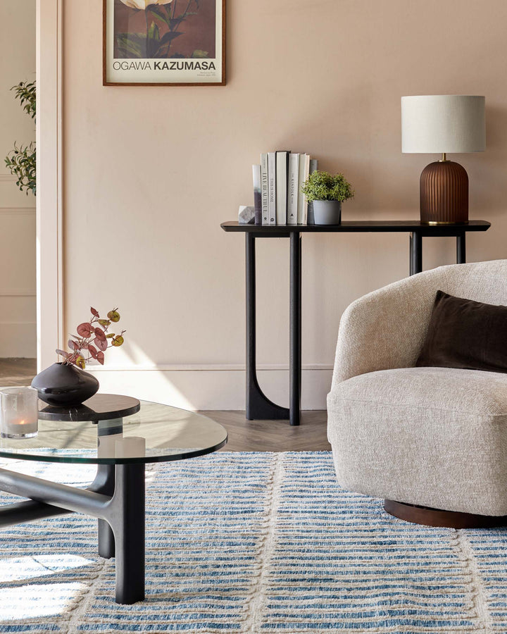 Living room furniture including a glass coffee table, side table with lamp, chair, and a patterned blue rug.