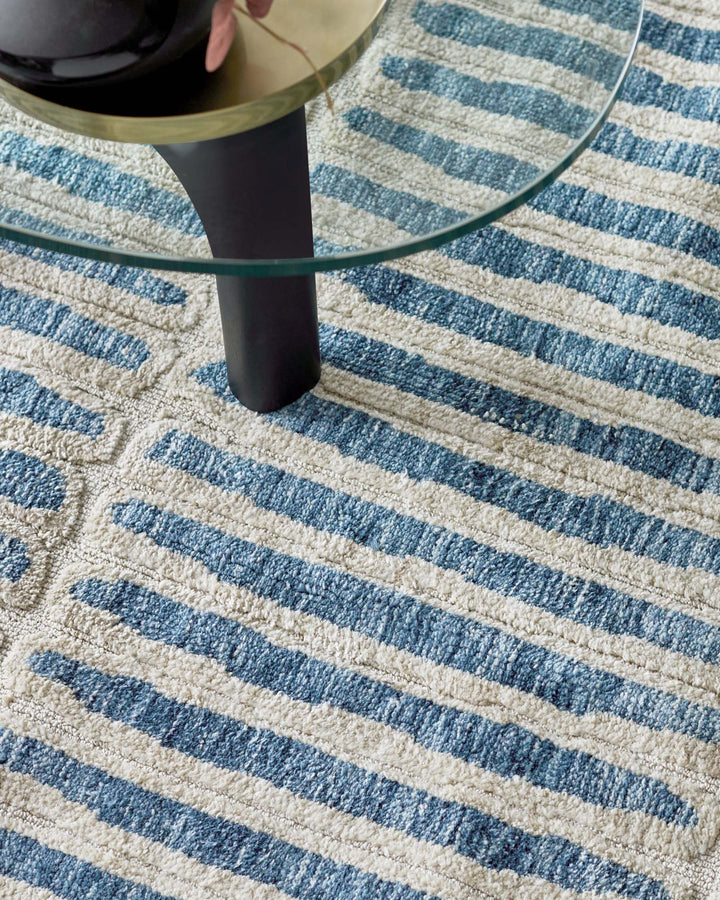 A round glass coffee table with a black, cylindrical base sits on a striped blue and cream rug.