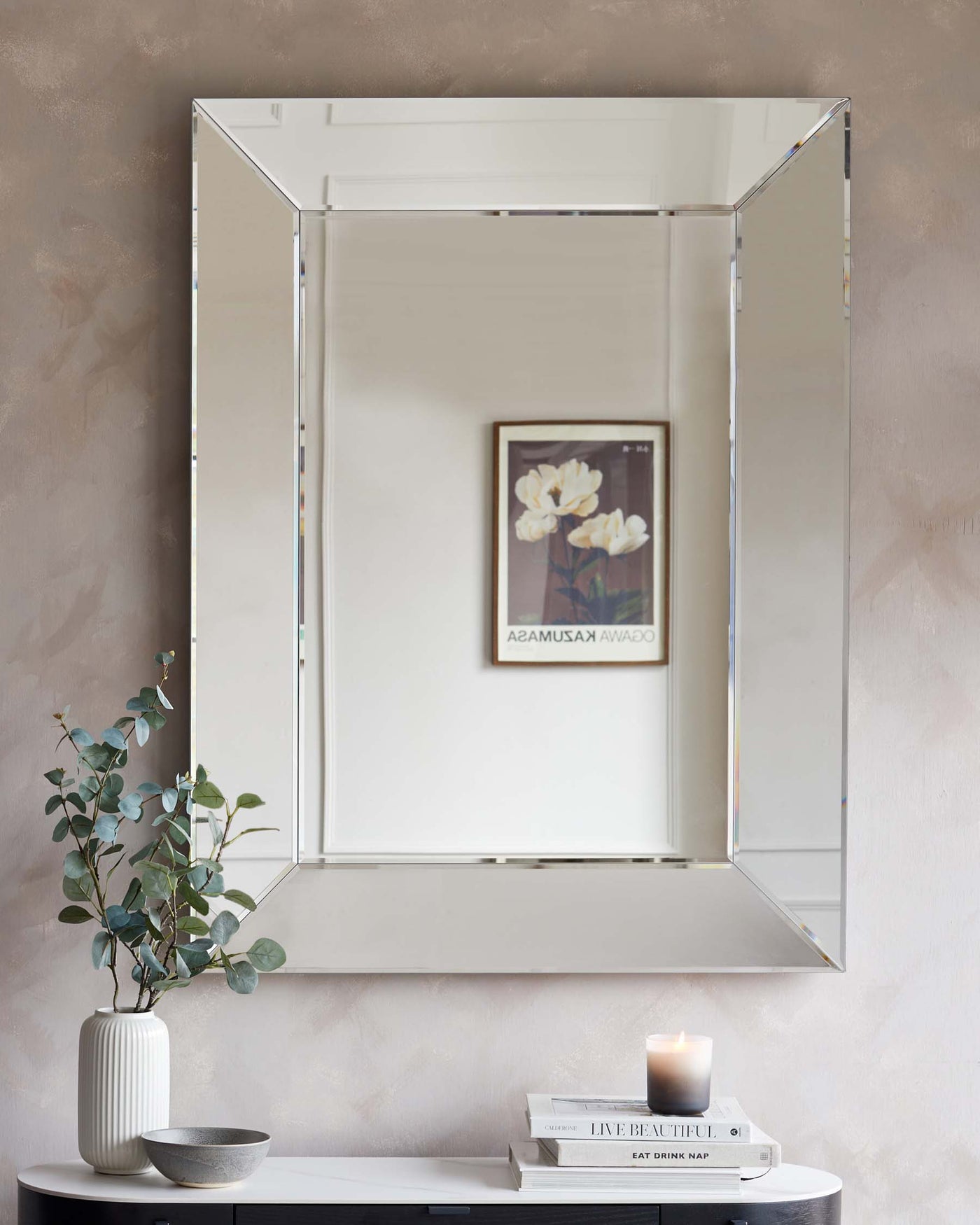Elegant rectangular bevelled mirror with a reflective frame, mounted above a modern minimalist black console table featuring books, a candle, a ceramic bowl, and a white vase with eucalyptus branches.
