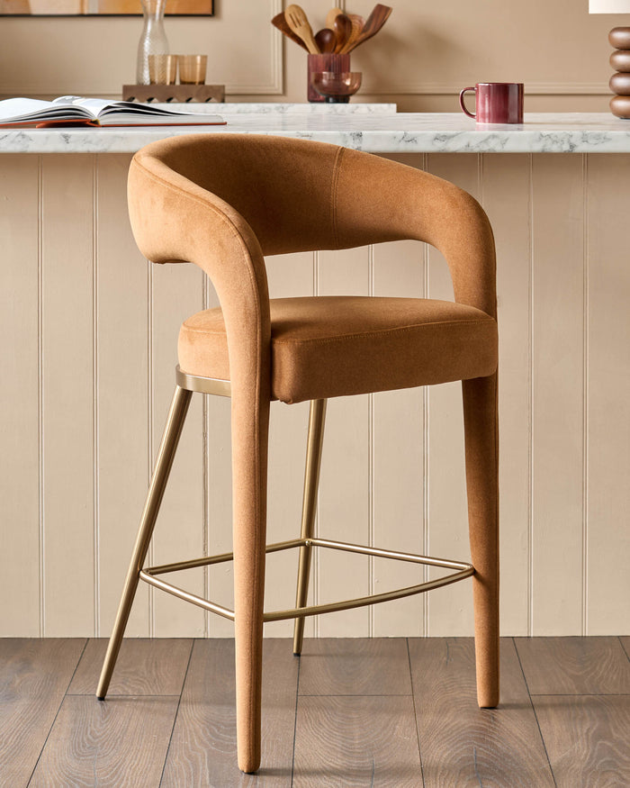 A stylish tan bar stool with a curved back and gold-coloured metal legs sits on a wooden floor in front of a neutral wall.
