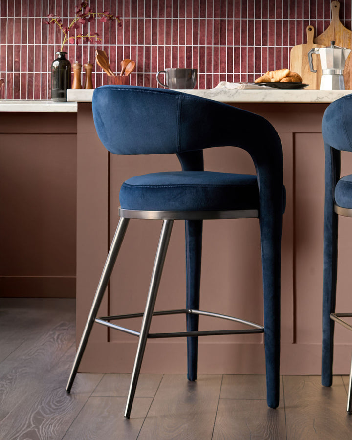 Rhett bar stool in dark navy velvet with curved backrest and brushed metal legs, shown at kitchen island.