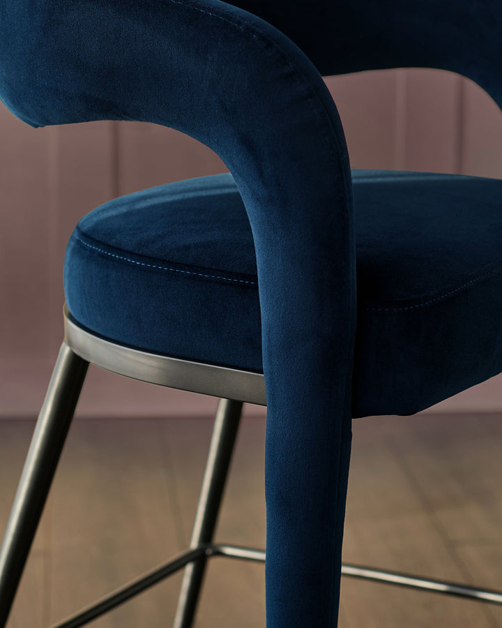 Rhett bar stool in dark navy velvet with curved backrest and brushed metal frame, side angle view.