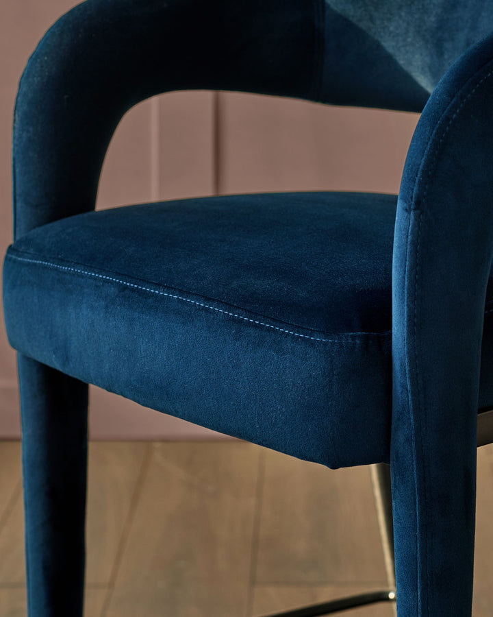 Dark navy velvet bar stool with curved backrest and wooden legs, angled side view showing upholstered seat and armrests.