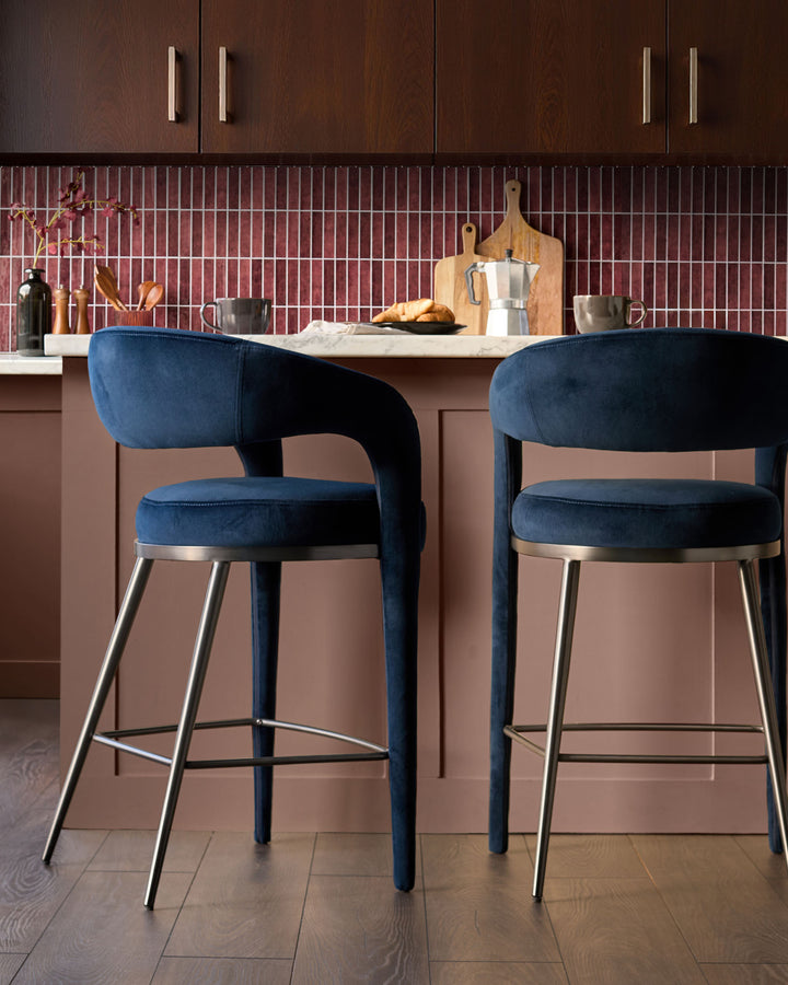 Rhett bar stool in dark navy velvet with curved backrest and brushed metal footrest, shown in kitchen setting.